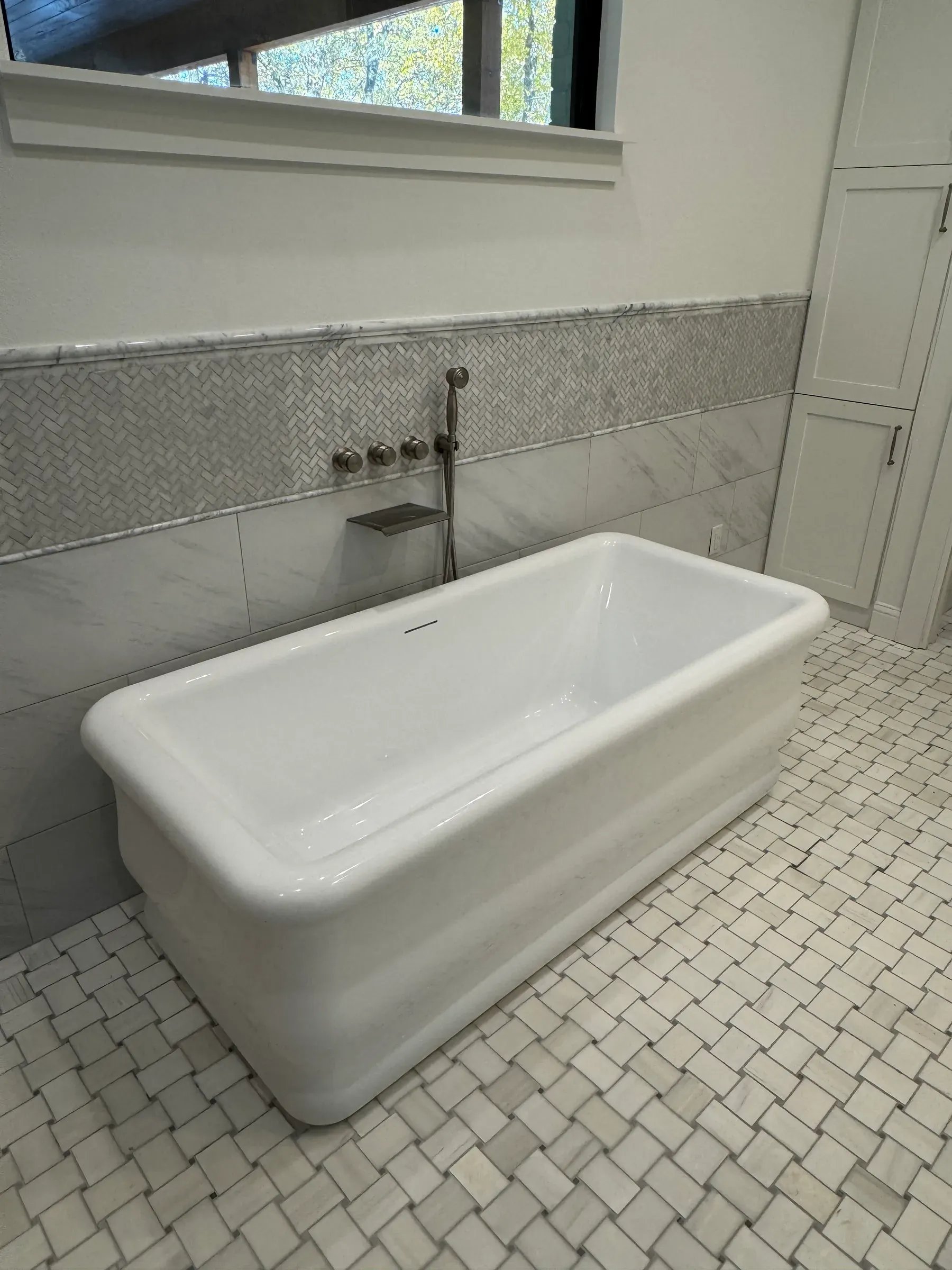 Freestanding soaking tub on a basketweave tile floor with a wall-mounted tub filler